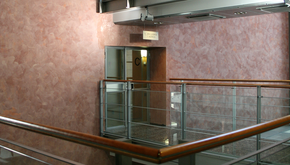 Rakennuksen aulakäytävä ja kerroksen ovi. Taustalla punertavaa seinää.