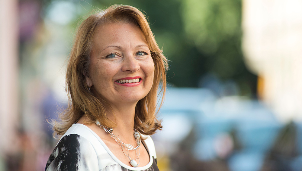 Päivi Sillanaukee. Nainen seisoo kadulla ja hymyilee. Taustalla autoja ja puita.