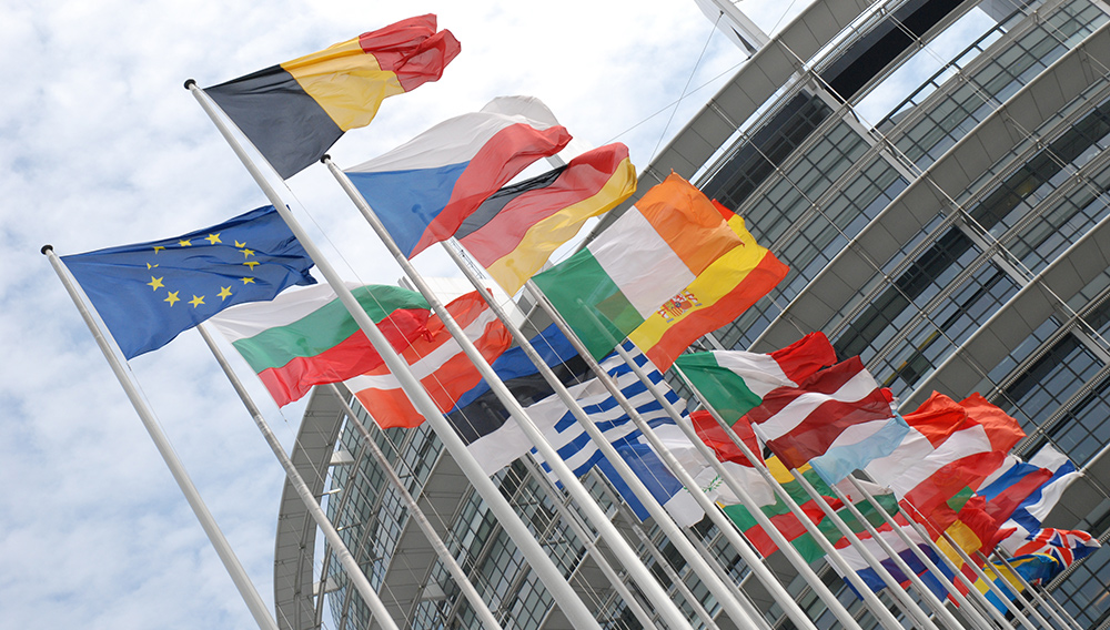 Useita Euroopan maiden lippuja sekä EU:n lippu liehuvat lipputangoissa. Taustalla näkyy harmaa lasinen rakennus.