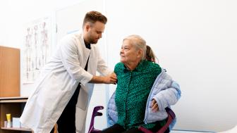 Kuvassa lääkäri auttaa vastaanotolla tuolille istahtavalta potilaalta takkia pois päältä. Kuva on kuvituskuva.