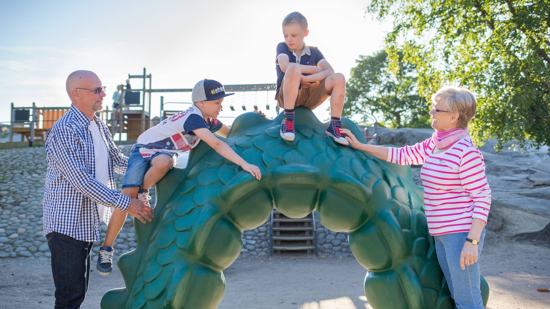 Kuvassa yksi lapsi istuu vihreän kiipeilytelineen päällä, yksi lapsi on kiipeämässä ylös ja häntä auttaa aikuinen. Toinen aikuinen on kiipeilytelineen toisella puolella.